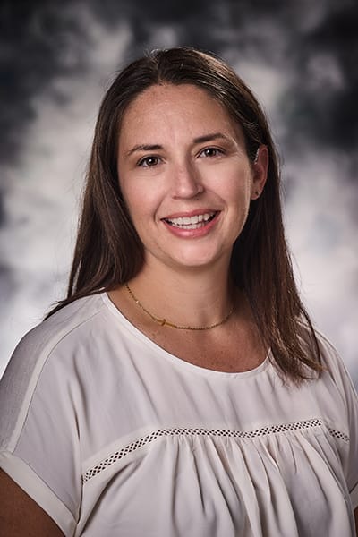 Woman with straight, shoulder-length brown hair wearing a white top, smiling in front of a studio gray background.