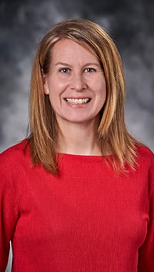 Woman with straight, shoulder-length blonde hair wearing a red top, smiling in front of a studio gray background.