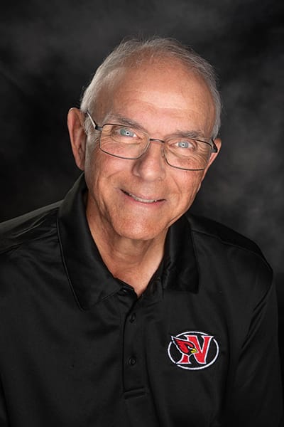 Man with white hair and glasses, wearing a black polo, smiling in front of a studio gray background.