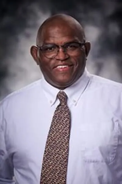 Portrait of a smiling man wearing glasses, a white button-down shirt, and a patterned tie, posed in front of a neutral studio background.